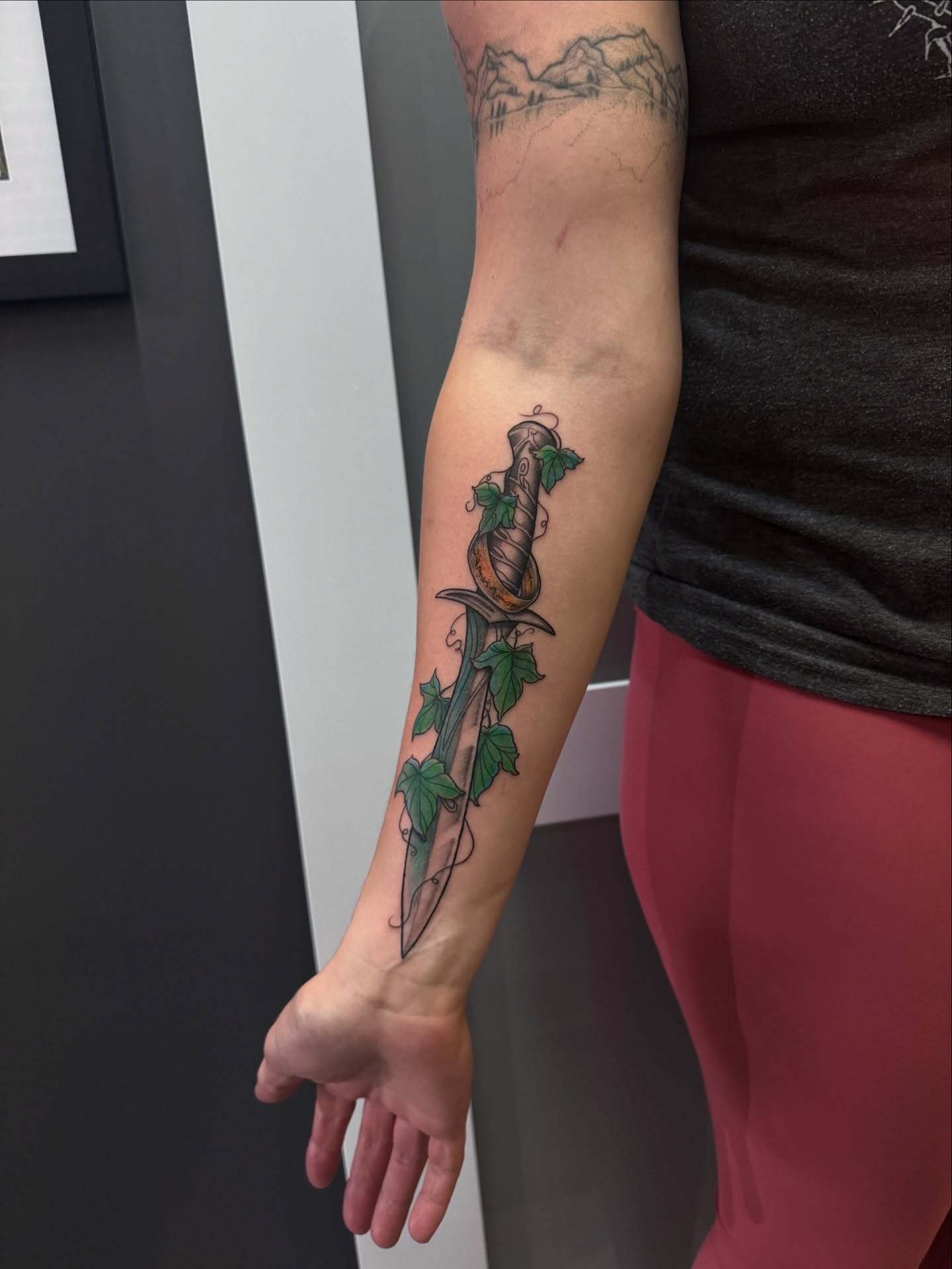 Custom illustrative forearm tattoo of Bilbo/Frodo's Sting Sword with the One Ring and green ivy vines, featuring clean linework and soft color shading by AJ at Poking Hazard in Edmonton, AB.