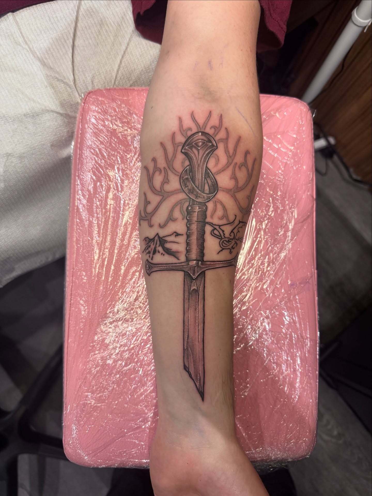 Custom black and grey illustrative forearm tattoo of the shards of Narsil broken sword with the One Ring, the White Tree of Gondor, and a small dragon, created by AJ at Poking Hazard in Edmonton, AB.
